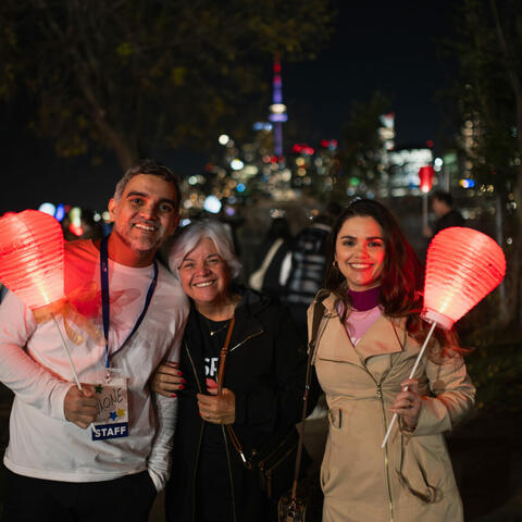 Three people standing with red glowing lanterns at night standing in front of a blurred CN tower and cityscape far in the background.
