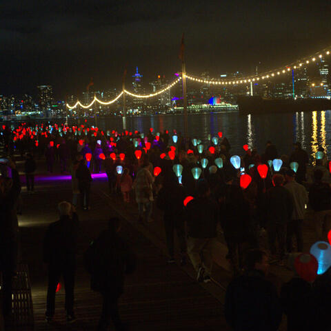 A large group of people walking at night with lit lanterns by a water dock